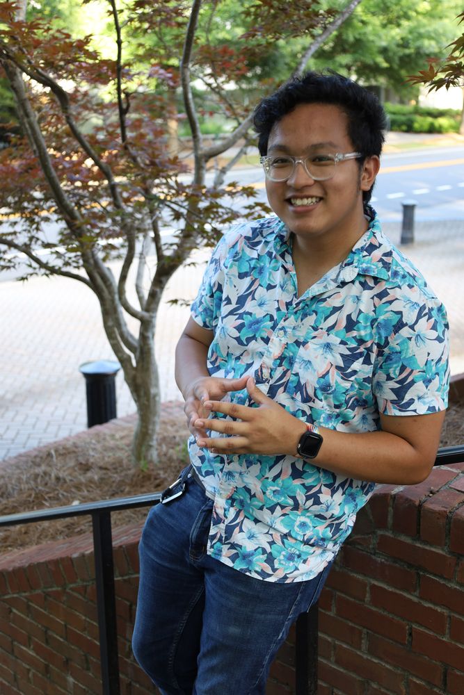 Man in a floral shirt leaning on a brick wall under a Japanese maple