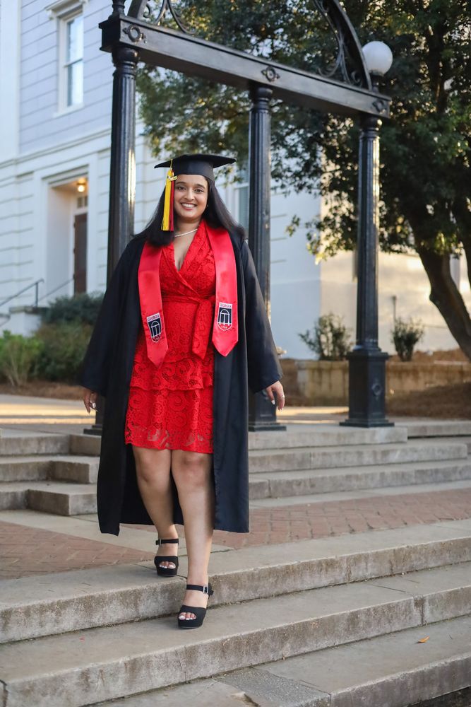 Graduate in a red dress, cap and gown walking down campus steps