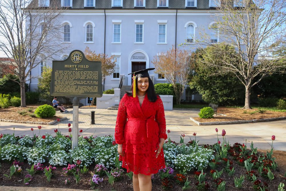 Graduate in a red dress and cap standing in a campus garden