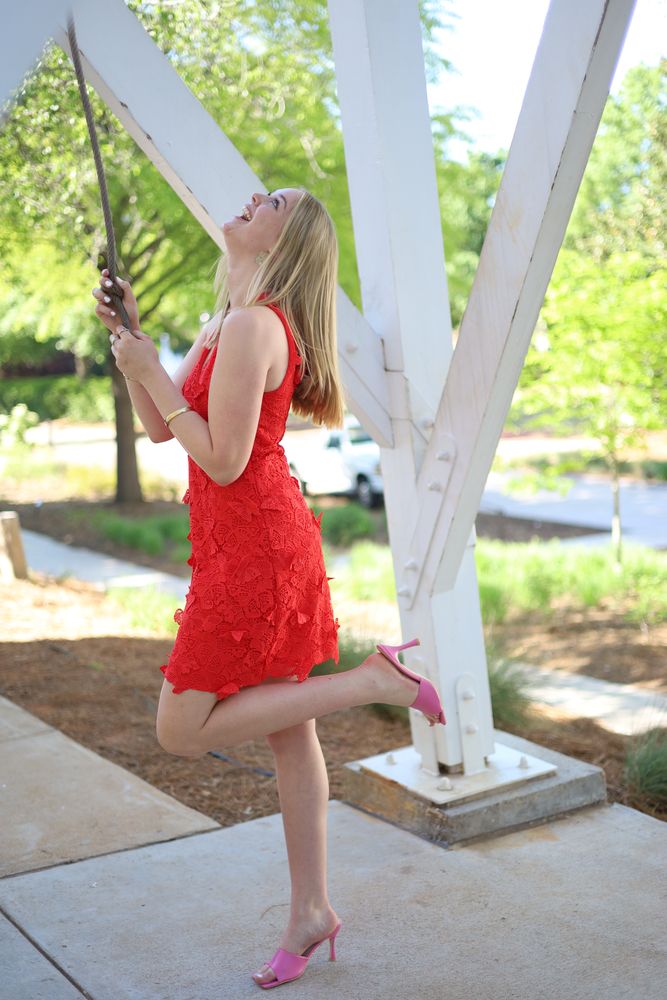 Graduate in a red dress kicking a heel under a white trellis