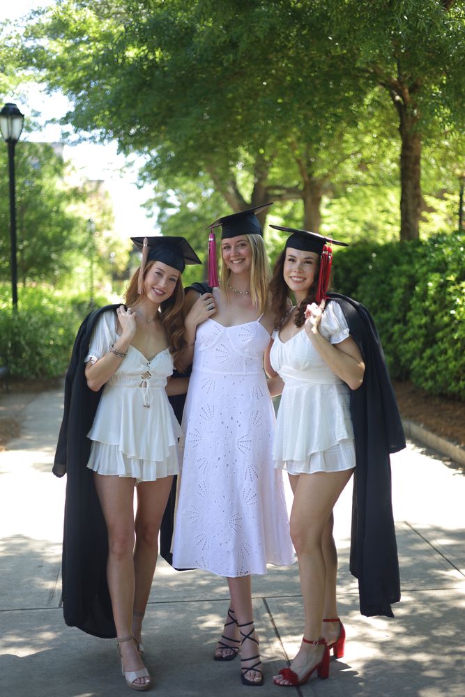 Three graduates in caps and white dresses on a tree-lined path