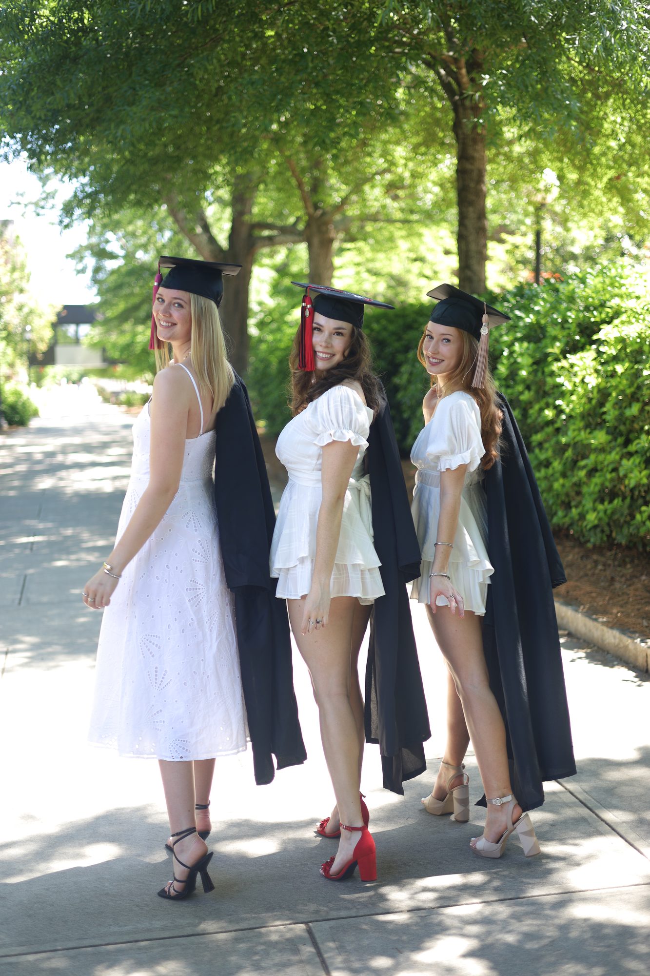 Three graduates in caps and white dresses on a tree-lined path