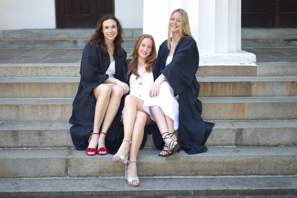 Three graduates in gowns seated on the Chapel steps, smiling at the camera